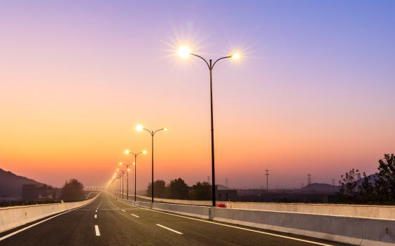 Imagem de uma estrada, com luminária LED acesas no separados das faixas de rodagem. O céu, em tons rosa, indica-nos que é fim do dia.