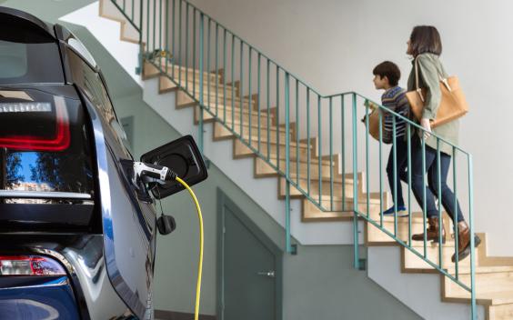 Imagem composta por um carro elétrico ligado a um carregador e, ao fundo, uma mulher uma criança a sobem umas escadas.